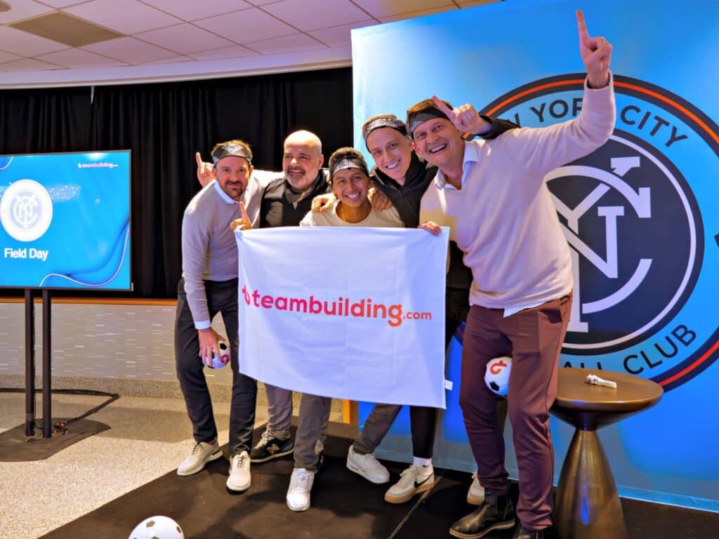 Men on a stage smiling and holding hands up in victory while holding a teambuilding.com banner